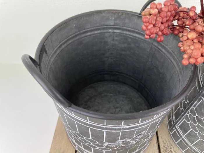 En grå metalspand med et geometrisk hvidt mønster, placeret på en træoverflade, med en klynge røde bær ved siden af.