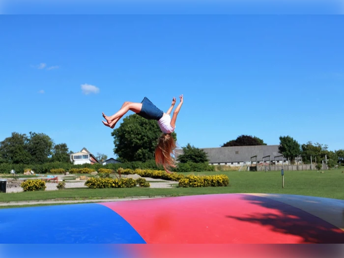 En pige i denimnederdel og hvid skjorte laver en baglæns salto i luften på en farverig udendørs hoppeborg med grønt græs og bygninger i baggrunden.