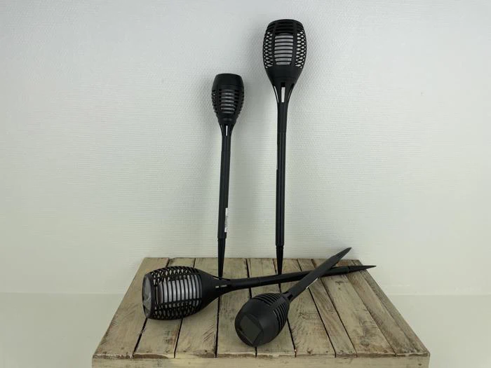 Tre sorte solcelle-havelamper står på en træoverflade, mens en fjerde fakkel ligger vandret foran dem. Baggrunden er en almindelig hvid væg.