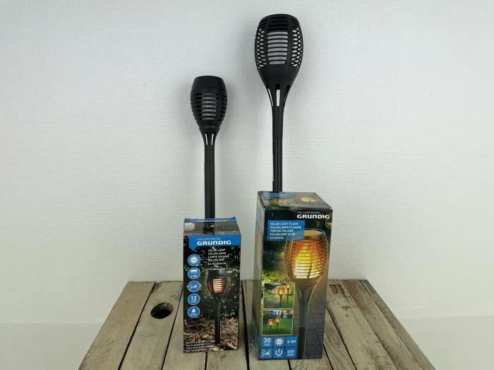 To sorte solcellelamper i fakkelstil står på en træoverflade ved siden af hver sin emballage, der viser produktegenskaber og en oplyst udendørs scene.