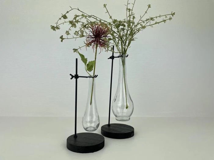To klare glasvaser med tynde metalstativer holder minimalistiske arrangementer af sarte blomster og grønt mod en almindelig hvid baggrund.
