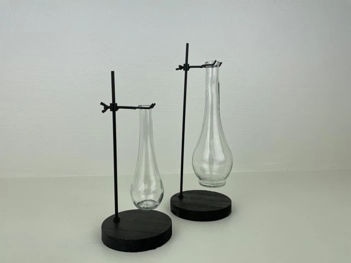To glasvaser i forskellige størrelser holdes oprejst af metalstativer med runde, sorte baser og står på en hvid flade mod en almindelig hvid baggrund.