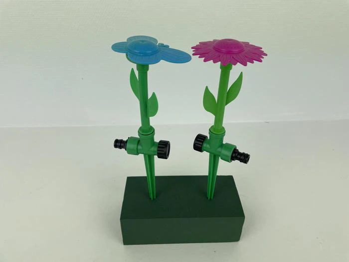 To farverige blomsterformede havesprinkler i plast, en blå og en lilla, er fastgjort til grønne stængler på en grøn rektangulær base.
