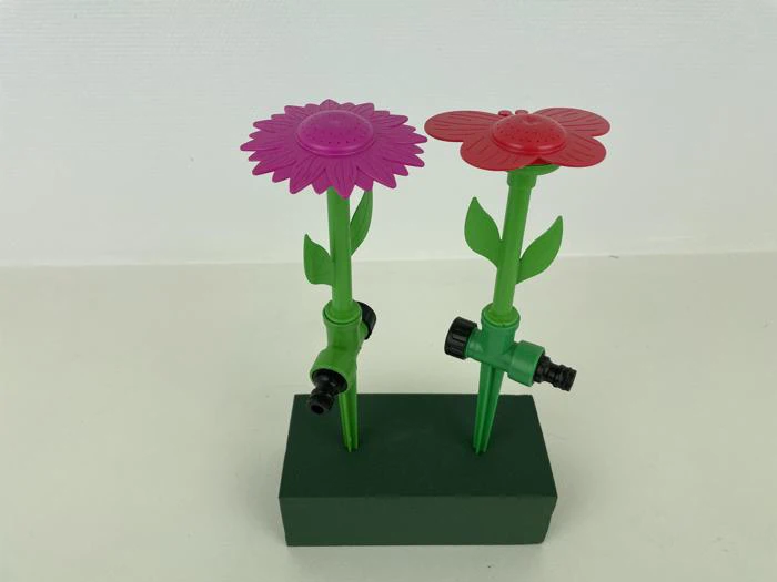 To blomsterformede haveslanger af plast, en lyserød og en rød, er monteret på grønne stængler med stik og står oprejst i en grøn skumblok.