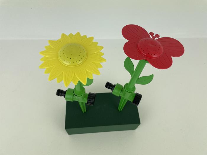 To farverige havesprøjter formet som en gul solsikke og en rød sommerfugl, med grønne stængler og slangekoblinger, står på en rektangulær grøn base.