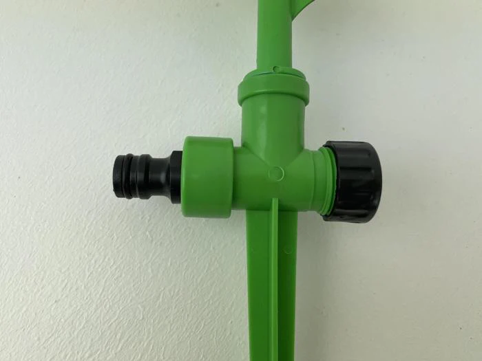 Nærbillede af en grøn plastikhavesprinkler med sort slangestuds og justeringsknap, placeret på en hvid flade.