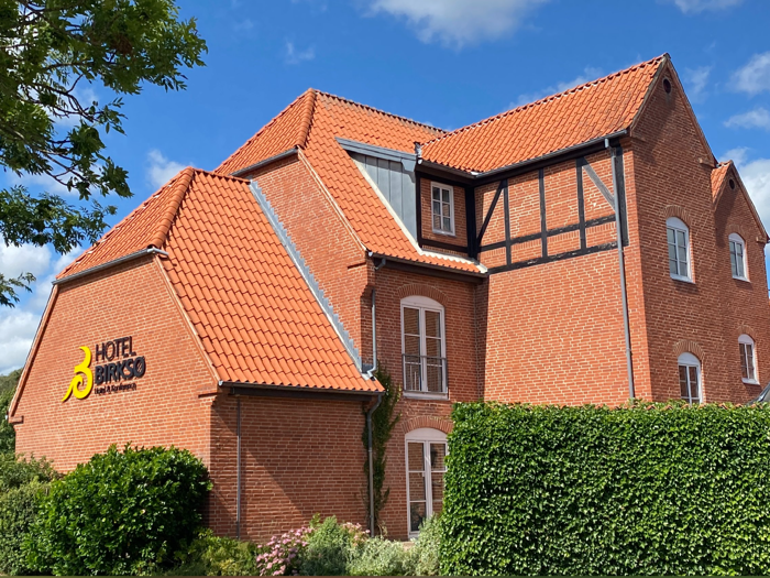 En rød murstensbygning med rødt tegltag, grønne hække og det gule Hotel Birke-skilt på væggen under en blå himmel med nogle skyer.
