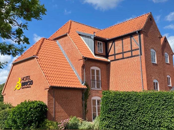 En rød murstensbygning med rødt tegltag, grønne hække og det gule Hotel Birke-skilt på væggen under en blå himmel med nogle skyer.