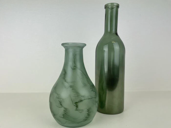 To grønne glasflasker står på en hvid flade; den ene er kort og rund med en bred hals, den anden er høj og slank med en smal hals.