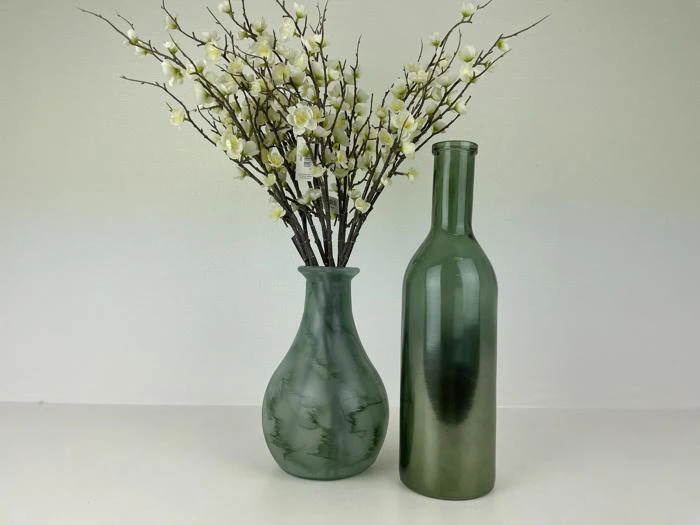 To grønne glasvaser: den ene med hvide kunstige blomster og grene, den anden tom, placeret side om side mod en almindelig lys baggrund.