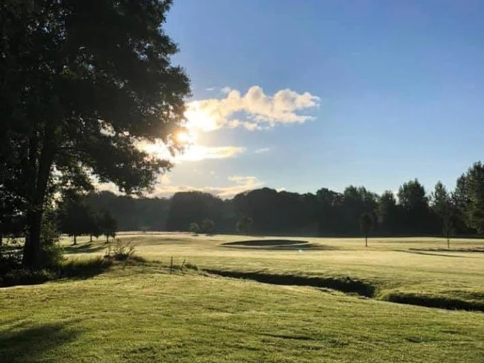 En solbeskinnet golfbane med grønt græs, sandbunker og træer rundt om kanten under en klar blå himmel med nogle skyer.