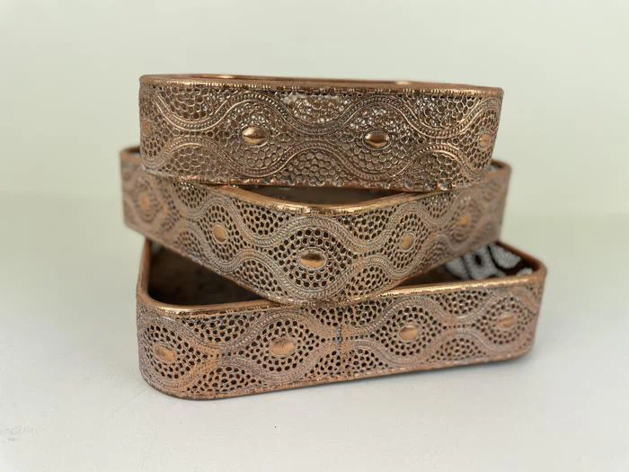 Tre udsmykkede, rektangulære kobberarmbånd med indviklede mønstre og ophøjede ovale design er stablet på en hvid overflade mod en lys baggrund.