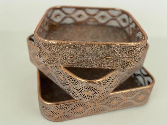 Tre sirligt mønstrede, rektangulære bronzebakker er stablet ujævnt mod en lys baggrund.