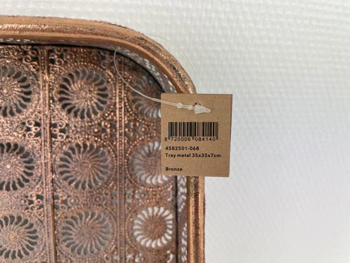 Nærbillede af en dekorativ metalbakke i bronze med blomstermønster og et mærke, hvor der står Bakke metal 35x35x7cm, Bronze.