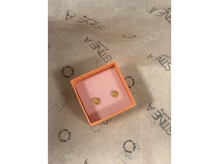 Et par små guldørestikker i et lyserødt smykkeskrin på hvidt silkepapir med ordet STINE A.