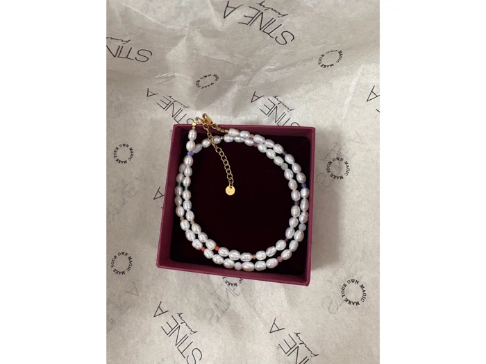 En dobbeltsnoet perlekæde med guldlås og charm, præsenteret i en rødbrun æske på mærket silkepapir med "STINE A" skrevet på.