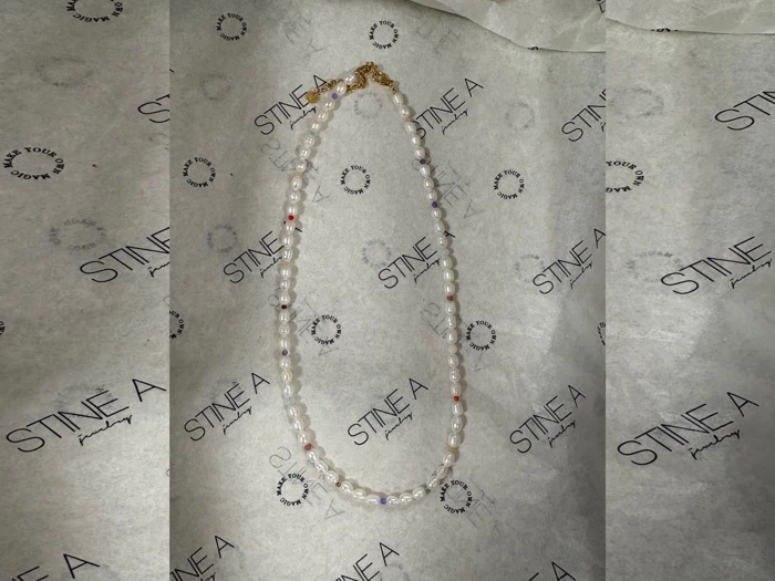 En perlekæde med hvide perler og små farvede perler vises på STINE A smykkepapir.