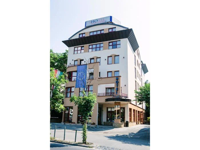 En moderne hotelbygning i flere etager med store vinduer, et blåt hotelskilt og flag udenfor, omgivet af træer på en solrig dag.