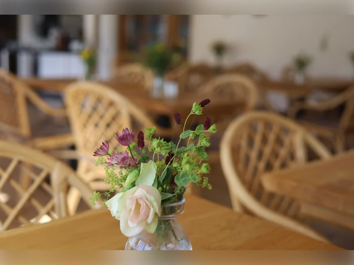 En lille buket blomster i en glasvase står på et træbord i en café med kurvestole og flere borde i den slørede baggrund.