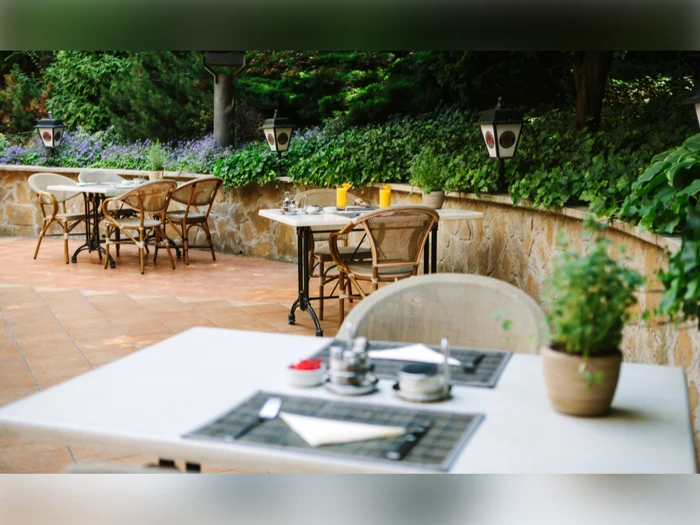Udendørs restaurantterrasse med borde og stole, dækkeservietter til spisning, potteplanter, grønne planter og dekorative lanterner langs en stenmur.