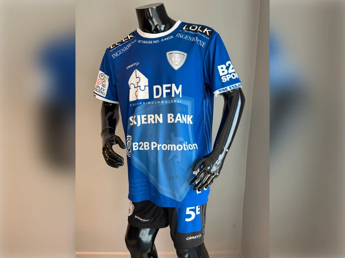 En sort mannequin iført en blå fodboldtrøje med forskellige sponsorlogoer og sorte shorts, der står indendørs foran en ensfarvet baggrund.