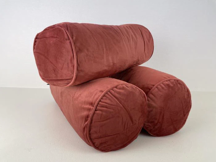 To cylindriske, rustfarvede fløjlspuder stablet i L-form mod en ensfarvet hvid baggrund.