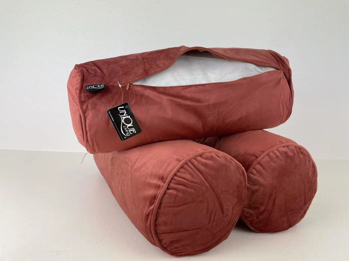 En rødbrun bolsterpude med lynlåsåbning, der viser hvidt fyld indeni, hviler på en hvid overflade. Puden har to cylindriske ender og et sort mærke med påskriften UNIQUE.