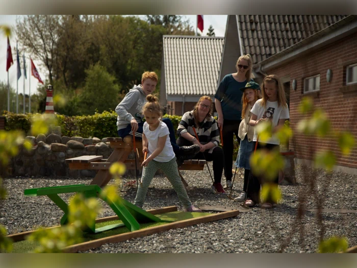 En ung pige spiller minigolf udenfor, mens fem personer – både børn og voksne – ser på og smiler i baggrunden på en solskinsdag.