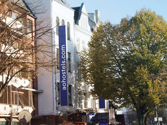 En hvid bygning med et lodret blåt skilt, hvor der står aohostels.com, omgivet af træer og nabobutikker på en solrig dag.