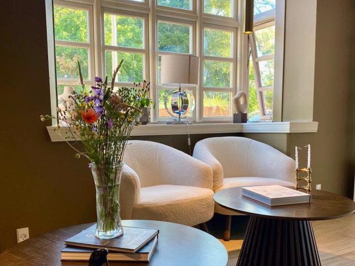 Et hyggeligt opholdsområde med to hvide lænestole ved et vindue, en vase med blomster på et bord, bøger og blødt naturligt lys, der strømmer ind.