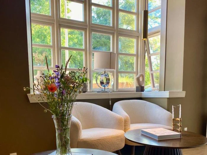 Et hyggeligt opholdsområde med to hvide stole, et rundt bord, en vase med vilde blomster, bøger og moderne indretning ved store, solbeskinnede vinduer med udsigt til grønne træer udenfor.