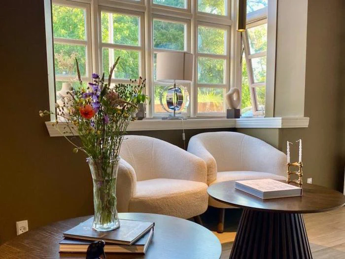 Et hyggeligt siddeområde med to hvide stole ved et vindue, en vase med blomster på et bord, bøger og moderne indretning i et godt oplyst rum.