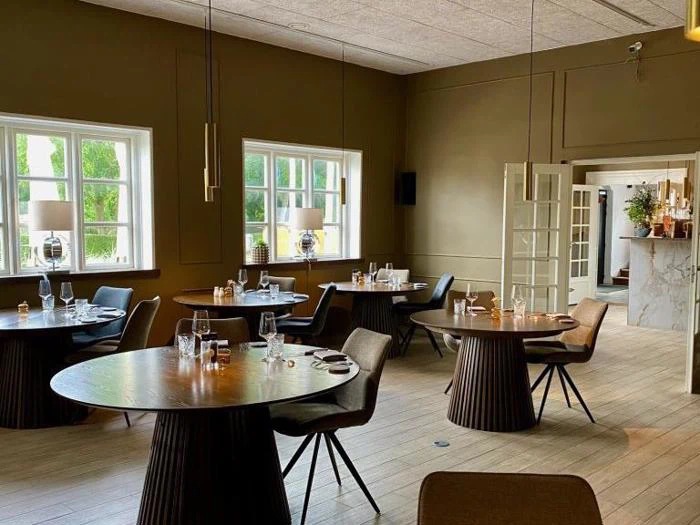 En moderne restaurantspisestue med runde træborde, dækket til gæster, og store vinduer, der lukker naturligt lys ind. Neutrale toner og minimalistisk indretning skaber en hyggelig atmosfære.