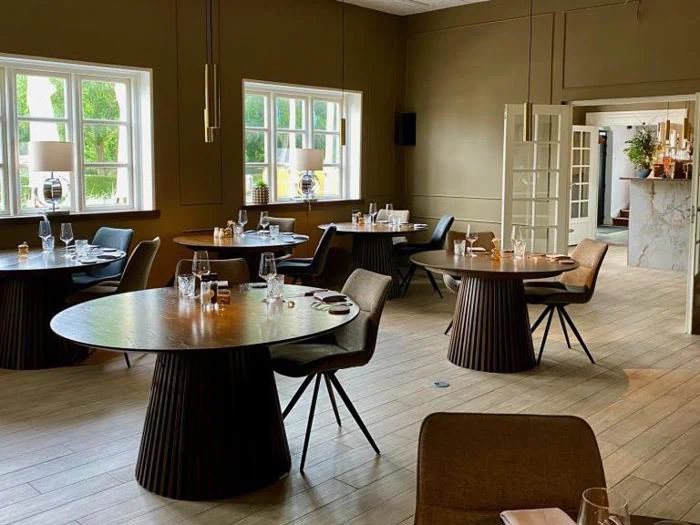 En moderne, elegant restaurant med runde træborde, pænt dækket med glas og bestik, brune stole og store vinduer, der lukker naturligt lys ind.