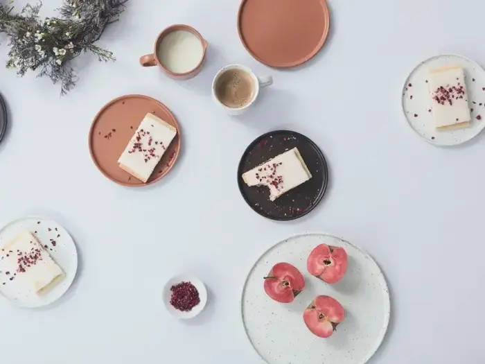 Fem tallerkener med kagestykker, en tallerken med fire æbler, krus med kaffe, en lille skål med frø og en buket blomster arrangeret på en hvid bordplade.