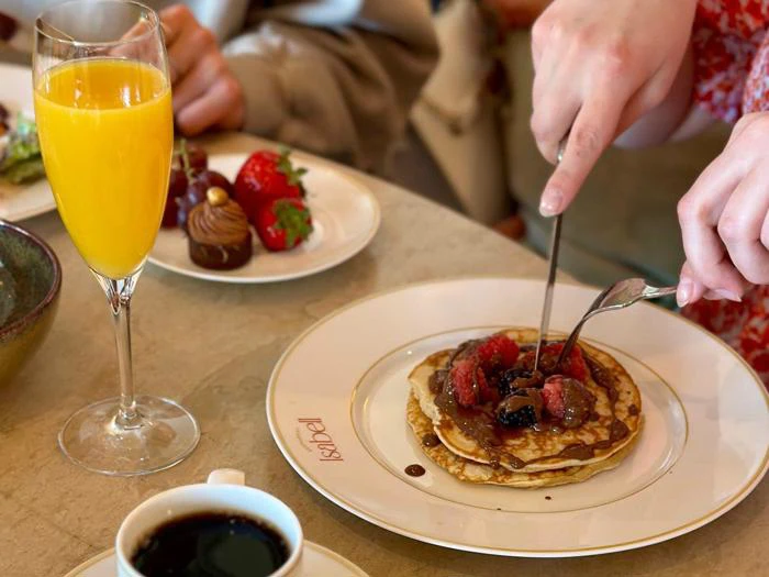 En person skærer i pandekager toppet med bær og chokoladesauce ved siden af et glas appelsinjuice, kaffe og jordbær med chokoladeovertræk.