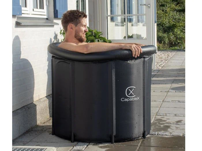 En mand slapper af i et lille, opretstående sort oppusteligt badekar med CapableX-logoet, som er placeret udendørs på en stenterrasse i nærheden af en hvid bygning.
