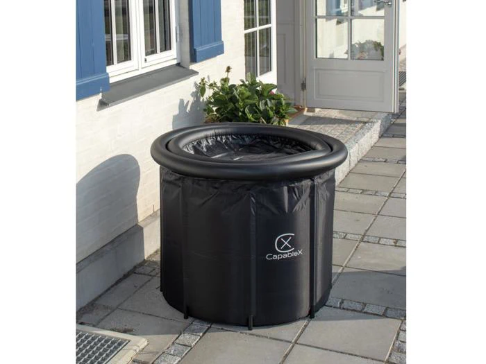 Et lille, rundt, sort oppusteligt badekar med logoet Capablex placeret på en flisebelagt terrasse uden for et hvidt hus med blå vinduesrammer og en åben dør.