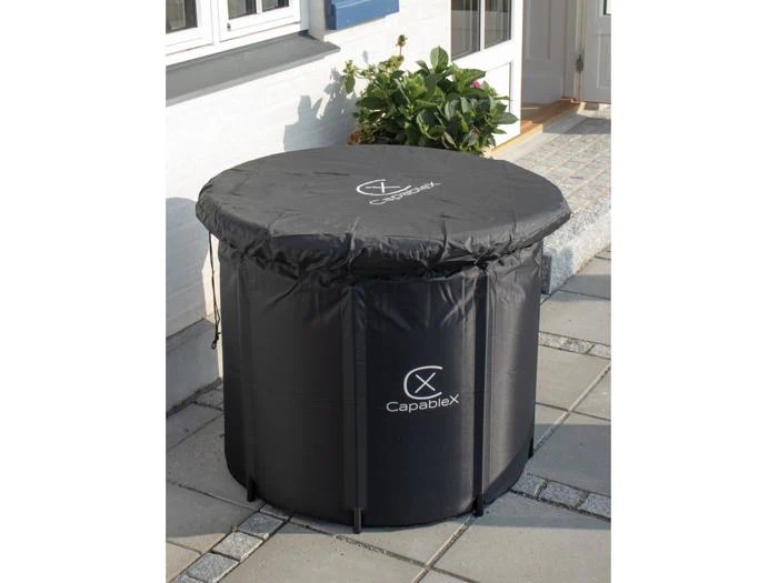 En rund, sort Capablex udendørs opbevaringsbeholder med monteret låg er placeret på en flisebelagt terrasse i nærheden af et hvidt hus og en grøn potteplante.