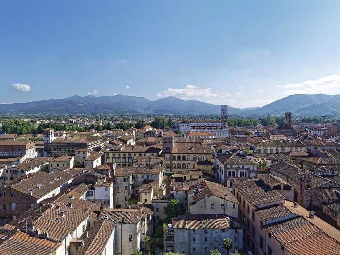 Luftfoto af en historisk italiensk by med røde tegltage, omgivet af grønne bakker og bjerge under en klar blå himmel.
