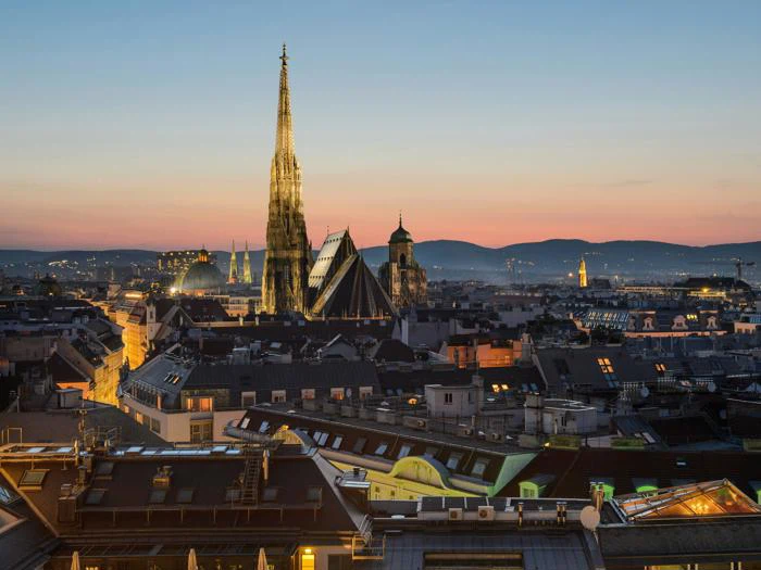 Et bybillede af Wien ved solnedgang med Stephansdom med sit høje spir og farverige tag, omgivet af historiske bygninger og blødt oplyst af aftenhimlen.