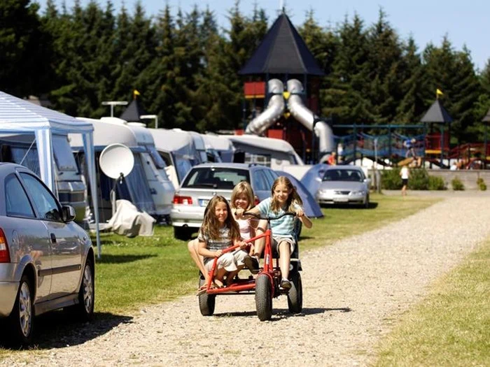 Tre børn kører på en gokart på en grussti på en campingplads med telte, biler og en legeplads med rutsjebaner i baggrunden.