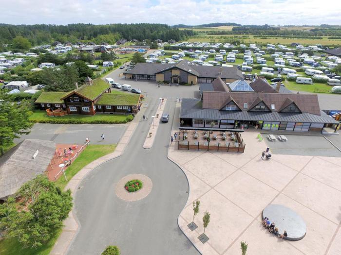 Luftfoto af en stor campingplads med mange parkerede autocampere, omkringliggende bygninger, stier og grønne områder på en delvis overskyet dag.