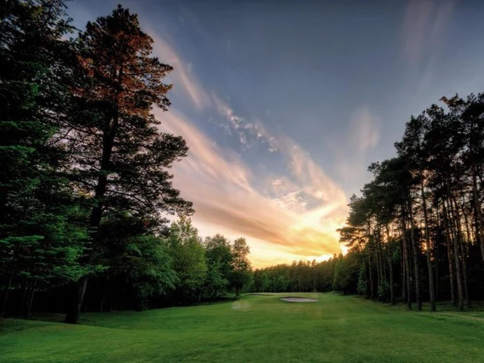 En naturskøn golfbane ved solnedgang med levende skyer på himlen, frodigt grønt græs og høje træer, der afgrænser fairwayen.