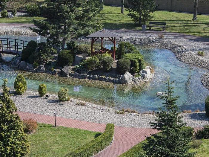 Et lille lysthus står på en klippeø omgivet af klart vand, træer og buske i en anlagt park med buede stier og en træbro.