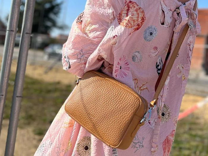 En person i en pink kjole med muslingemønster bærer en lysebrun crossbody-taske med struktur udendørs.