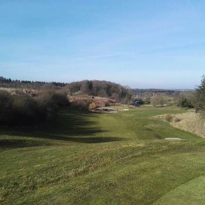 En græsklædt golfbane med bølgende bakker, sandfælder og træer under en klar blå himmel på en solrig dag.