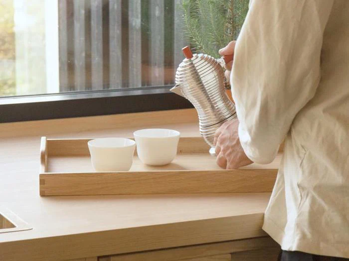 En person skænker te fra en sølvtekande i hvide kopper på en træbakke nær et vindue i et lyst, minimalistisk køkken.