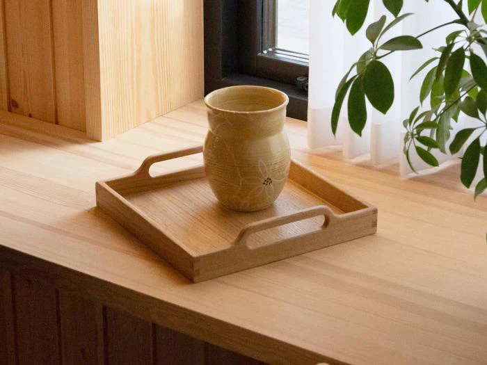 En keramisk kop står på en træbakke, der er placeret på en lys træoverflade nær et vindue med et gennemsigtigt gardin og en grøn plante.