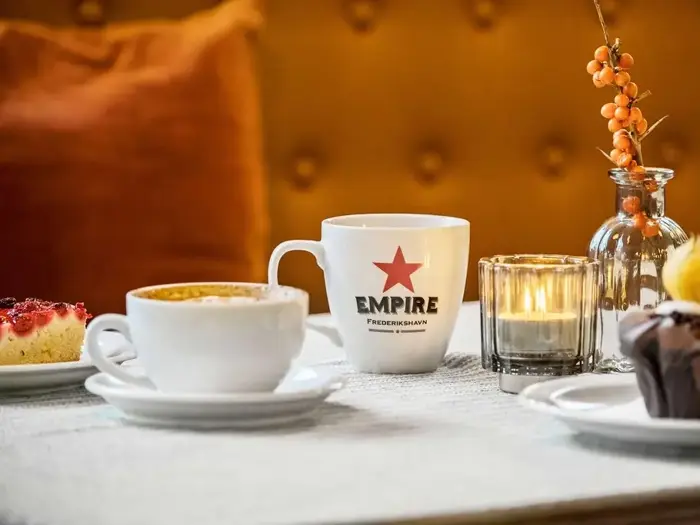 To kaffekopper, et stykke kage, en muffin, et tændt stearinlys og en lille vase med orange bær på et bord med en struktureret dug.